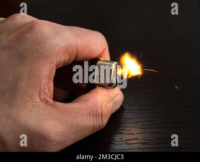 Nahaufnahme zu einer männlichen Hand, die ein gelbes Feuerzeug mit Flamme über einem dunklen Holztisch im Hintergrund hält Stockfoto
