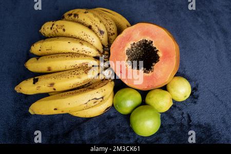 Draufsicht auf eine frisch geschnittene Orangenpapaya mit Samen im Inneren, eine Sommersprosse Bananen und drei grüne Zitronen auf blauem marineblauem und schwarzem Hintergrund. F Stockfoto