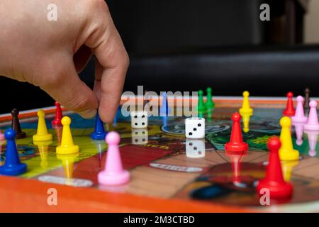 Männliche Hand mit einem blauen Bauern mit alten Würfeln und Farbenbauern über kolumbianischem Brettspiel mit dem Namen "Parqus" und Wohnzimmerhintergrund. Stockfoto