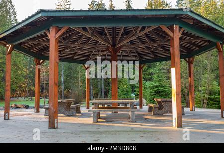 Holzpergola im Park mit Gartentischen und Stühlen im Schatten. Das hölzerne Sommerhaus befindet sich in einem grünen Park am Morgen. Niemand, selektiver Fokus Stockfoto