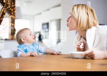 Mutter motiviert ihren kleinen Jungen, während sie ihn füttert und zu Hause im Hochstuhl am Esstisch sitzt. Einführungskonzept für feste Babynahrung. Stockfoto