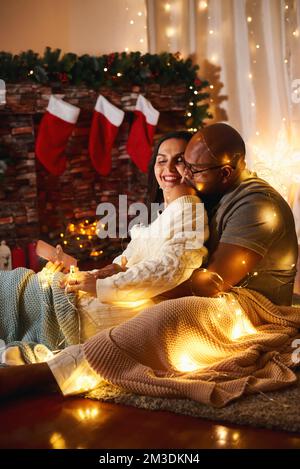 Ein glückliches, kaukasisches, afroamerikanisches Paar, das sich umarmt und in gemütlichen Decken mit Girlande vor dem Kamin sitzt, Geschenksocken zur Feier des Neujahrs C Stockfoto