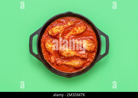 Draufsicht mit Hühnereintopf in einem eisernen Gusstopf Minimalist auf einem grünen Tisch. Köstlicher Fleischeintopf mit Hähnchenschenkeln, Tomatensoße und Gewürzen. Stockfoto