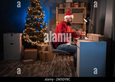 Junger Anfänger, bärtiger hindu-Unternehmer, kleiner Unternehmer im weihnachtsmann-Kostüm, müde, nachts im Home Office zu arbeiten, rote Schleife an del zu binden Stockfoto