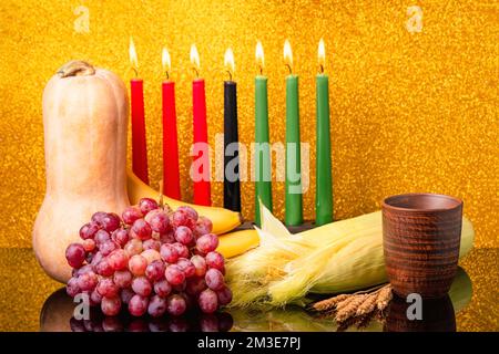 Kwanzaa Urlaubskonzept mit sieben Kerzen Rot, Schwarz und Grün, Kürbis, Schüssel, Mais und Obst auf einem leicht verschwommenen Bokeh-Hintergrund mit Reflektio Stockfoto