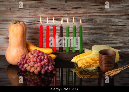 Kwanzaa-Urlaubskonzept mit traditionellen Kerzen, Kürbis, Mais, Trauben, Banane, Schüssel und Weizen mit Reflexion auf schwarzem Hintergrund Stockfoto