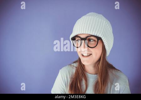 Ein junges Mädchen mit dicken, massiven Gläsern und Beanie vor lila Hintergrund Stockfoto