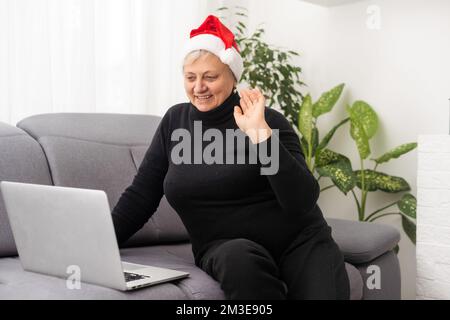 Lächelnde Seniorin mit Weihnachtsmannmütze zur Weihnachtszeit, die von zu Hause aus auf dem Laptop tippt und mit Familie oder Freunden in Kontakt kommt. Stockfoto