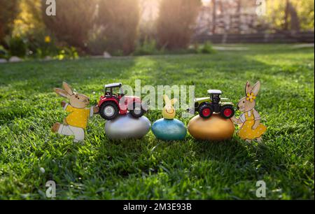 Modelle von Spielzeugmodellen von zwei Traktoren, bunten Eiern und osterhasen liegen auf dem Gras. Geschäftskonzept für Ostergrüße für Landwirtschaft und Stockfoto