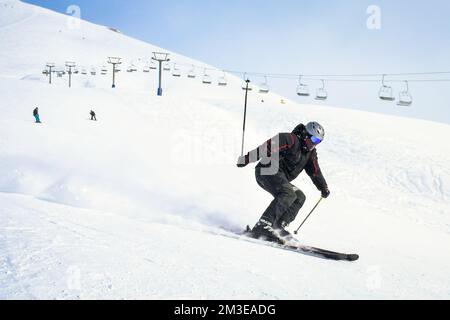 Coole männliche Skifahrer in Schwarz aus nächster Nähe fahren bergab und schnitzen rasanten, bedeckten Schnee mit Bergen und Skilift im Hintergrund Stockfoto