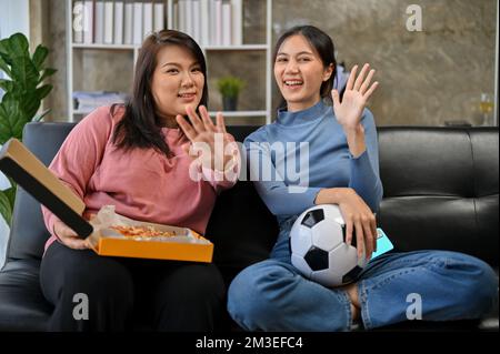 Zwei hübsche asiatische Freundinnen sitzen auf dem Sofa im Wohnzimmer, feiern und sehen sich zusammen ein Fußballspiel an. Stockfoto