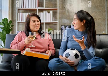 Zwei glückliche Millenials-asiatische Freundinnen sitzen auf dem Sofa und unterhalten sich, während sie sich zu Hause ein Fußballspiel ansehen. Stockfoto