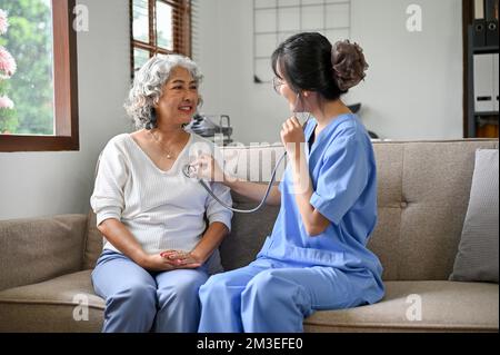 Professionelle asiatische Ärztin besucht ihre pensionierte Patientin zu Hause und prüft den Herzschlag mit dem Stethoskop. Stockfoto