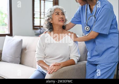 Eine junge Ärztin oder Krankenschwester, die während eines Besuchs zu Hause mit einer älteren Patientin kuschelt, spricht und beruhigt. Stockfoto