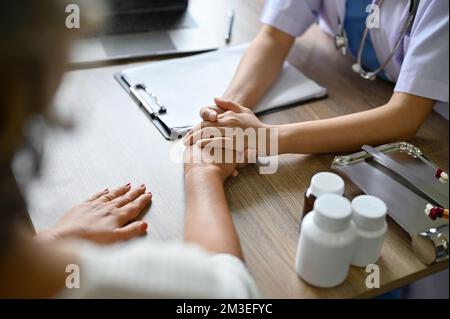 Eine Ärztin, die die Hände ihrer Patientin auf dem Tisch hält, während sie den Behandlungsplan bespricht. Zugeschnittenes Bild und Nahaufnahme Stockfoto