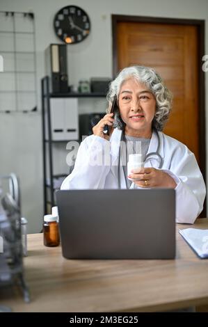 Porträt, professionelle asiatische Ärztin im Alter ruft jemanden an, telefoniert und trinkt Kaffee während der Arbeit in der Klinik. Stockfoto