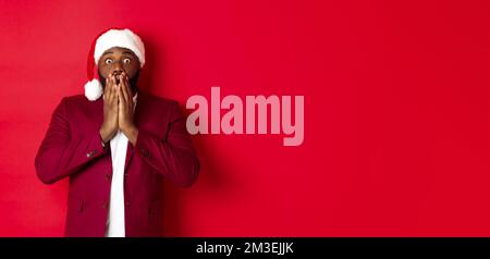 Weihnachts-, Party- und Feiertagskonzept. Schockiert afroamerikanischen Kerl in santa hat Reaktion sprachlos auf Werbung, stand vor rotem Hintergrund Stockfoto