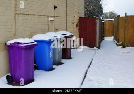 Eine Reihe von abfalleimern in verschiedenen Farben außerhalb eines Hauses bei schneebedeckten Winterwettern Stockfoto