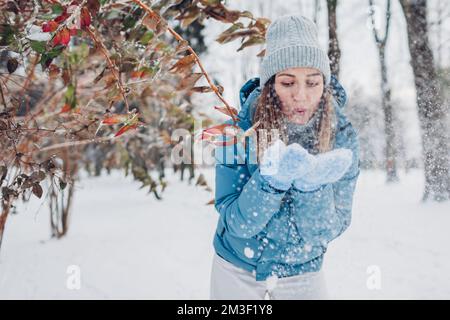 Glückliche junge Frau bläst Schnee aus den Händen, um Spaß im Winterwald zu haben. Mädchen mit blauem Mantel und Fäustlingen. Spielen im Freien bei kaltem Wetter Stockfoto