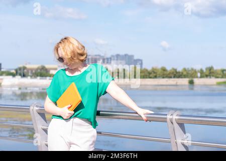 Eine lächelnde Bloggerin in einer Sonnenbrille, die ein Notebook oder Tablet in der Hand hält, am Ufer des Ufers. Fernunterricht online-pädagogisch Stockfoto