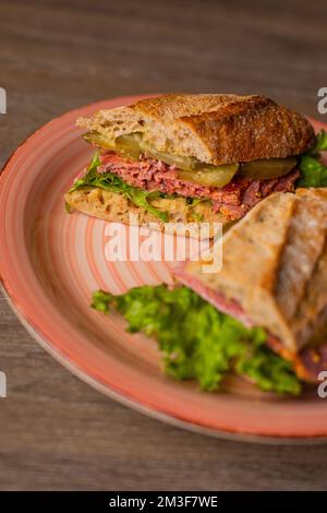 Vertikaler Schuss einer rosafarbenen Platte mit leckerem, knusprigem Sandwich mit Fleisch und Gurken, Petersilie, aufgeteilt in zwei Teile auf dem Tisch Stockfoto