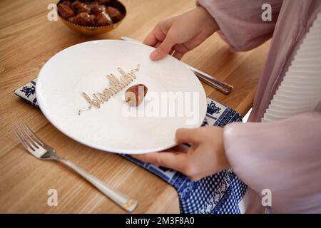 Muslimische Frauenhände servieren Tisch mit Besteck und Teller mit Datteln, Früchten und Inschrift bismillah. Eid al Fitr. Die Hände schließen sich Stockfoto