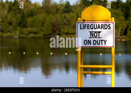 Hinter einem leeren Rettungsschwimmer-Stuhl mit einem Schild mit der Aufschrift RETTUNGSSCHWIMMER NICHT IM DIENST. See im Hintergrund mit Bäumen über dem See. Geringe Schärfentiefe Stockfoto