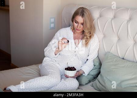 Schwangere Frau, die auf dem Bett sitzt und Kirschbeeren isst. Schöne Schwangerschaft. Blondes Model im Pyjama Stockfoto