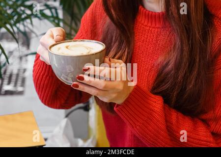Frauenhände mit roter Maniküre halten eine große Tasse Keramikkaffee. Duftender Cappuccino in einem Café. Stockfoto