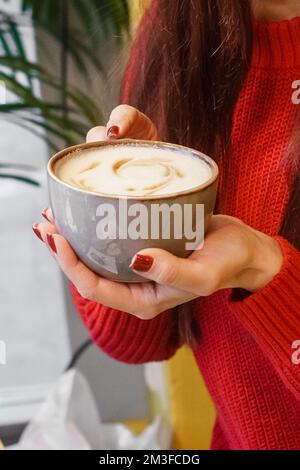 Frauenhände mit roter Maniküre halten eine große Tasse Keramikkaffee. Duftender Cappuccino in einem Café. Stockfoto