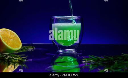 Nahaufnahme des Gießens eines grünen Cocktailgetränks in einem transparenten Glas, das auf der Theke steht. Clip. Die Bar ist mit grünen Blättern und oran dekoriert Stockfoto