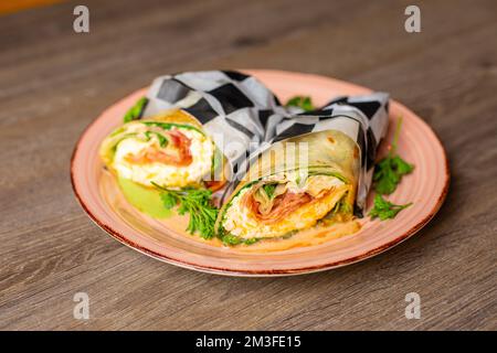 Leckere Speckbrötchen mit Grünzeug, Sauce und Schinken, verpackt in karierte Serviette, serviert auf einem Holztisch Stockfoto