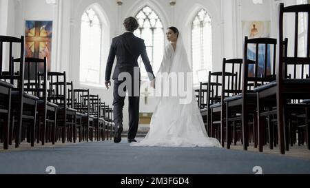 Braut und Bräutigam bei der Hochzeitszeremonie in der modernen Kirche. Aktion. Frau und Mann gehen zusammen auf den Altar zu, inmitten der leeren Sitzreihen Stockfoto