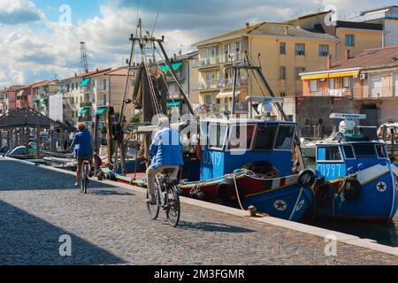 Ältere Radfahrer, Rückblick auf zwei ältere Radfahrer, die ihre Fahrräder entlang eines Kais in einem italienischen Fischereihafen, Chioggia, Veneto, Italien, fahren Stockfoto