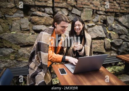 Positives, multiethnisches Paar in der Decke mit Laptop auf der Terrasse des Cafés, Stockbild Stockfoto