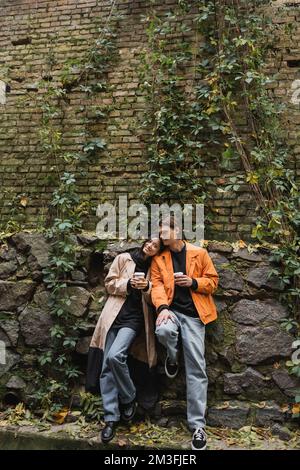 Lächelnde asiatische Frau im Trenchcoat, die in der Nähe ihres Freundes auf die Kamera schaut, mit Kaffee, um auf die urbane Straße zu gehen, Stockbild Stockfoto