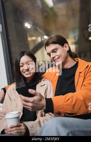 Fröhliches, multiethnisches Paar mit einem Getränk zum Mitnehmen mit dem Smartphone auf der Terrasse des Cafés, Stockbild Stockfoto