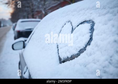 Herzform als Liebesschild an verschneiten Autofenstern. Im Winter können Sie auf einer schneebedeckten Straße parken. Stockfoto