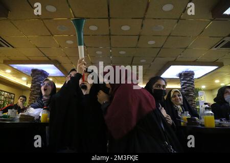 Teheran, Teheran, Iran. 15.. Dezember 2022. Getarnte iranische Fans reagieren, während einer von ihnen ein Horn hält, während er das Halbfinale der FIFA-Weltmeisterschaft Katar 2022 zwischen Frankreich und Marokko am 15. Dezember 2022 im Nakhlestan Café im Zentrum von Teheran, Iran, auf dem Bildschirm sieht. Fans aus dem Iran, dem Libanon, dem Jemen, Syrien und Palästina versammeln sich im Nakhlestan Cafe, das vom Owj, der Arts and Media Organization des Islamic Revolutionary Guard Corps (IRGS), betrieben wird und sich im Besitz der Organisation für Kunst und Medien befindet, um die marokkanische Nationalmannschaft zu unterstützen. Nach dem Sieg gegen Portugal in Katar am 10.. Dezember trugen die Marokkaner palästinensische Flaggen nach Katar Stockfoto