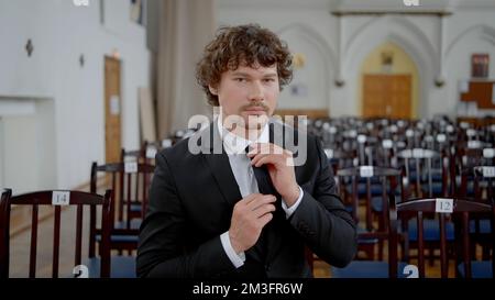 Junger Mann mit lockigem Haar in schwarz-weißem klassischem Anzug in leerer Kirche. Aktion. Der Bräutigam hat sich auf seine Hochzeit vorbereitet Stockfoto