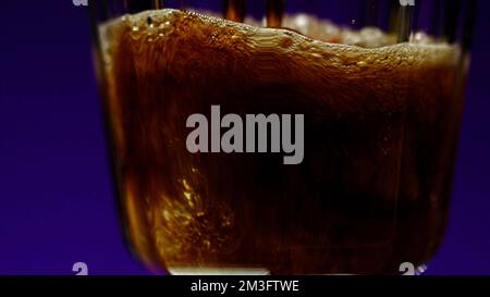 Ich gieße Cola mit Luftblasen in das Glas, Nahaufnahme. Clip. Cola Limonade in transparentem Glas auf einem bunten beleuchteten Wandhintergrund Stockfoto