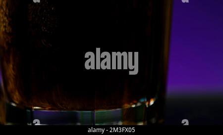 Ich gieße Cola mit Luftblasen in das Glas, Nahaufnahme. Clip. Cola Limonade in transparentem Glas auf einem bunten beleuchteten Wandhintergrund Stockfoto