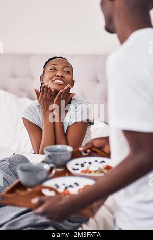 Ich habe den Jackpot geknackt, als ich bei dir eingezogen bin. Ein junger Mann bringt seiner Freundin Frühstück ins Bett. Stockfoto