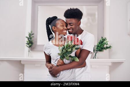 Ich will sie nur glücklich machen. Eine junge Frau, die einen Haufen roter Rosen hält, während sie mit ihrem Freund zu Hause steht. Stockfoto