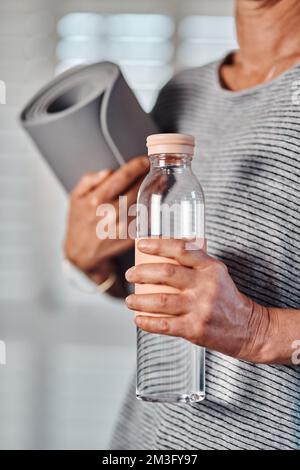 Meine Yoga-Essentials. Eine nicht wiedererkennbare, reife Frau, die eine Flasche Wasser und Yoga-Matte in der Hand hält, um ihre morgendliche Yoga-Session in einem zu beginnen Stockfoto