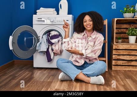Junge afroamerikanische Frau, die auf dem Boden sitzt und Wäsche wäscht, lächelt fröhlich mit Hand und Finger zur Seite zeigt Stockfoto