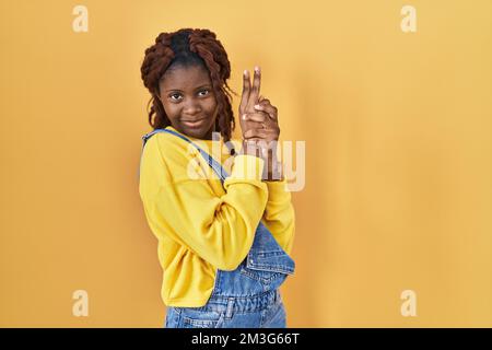 Afrikanische Frau, die über gelbem Hintergrund steht, symbolische Waffe mit Handgesten hält, tötende Schusswaffen spielt, wütendes Gesicht Stockfoto