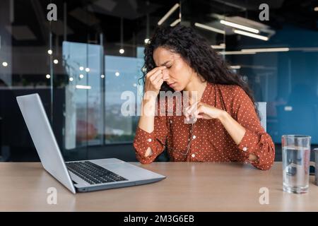 Übermüdete hispanische Frau bei der Arbeit, Geschäftsfrau mit Brille, die Hände am Kopf hält, hat starke Kopfschmerzen, arbeitet im Büro, sitzt am Tisch und benutzt ein Notebook bei der Arbeit. Stockfoto