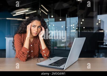 Übermüdete hispanische Frau bei der Arbeit, Geschäftsfrau mit Brille, die Hände am Kopf hält, hat starke Kopfschmerzen, arbeitet im Büro, sitzt am Tisch und benutzt ein Notebook bei der Arbeit. Stockfoto