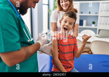 Ein junger Junge, der in einer Arztpraxis geimpft wird, zeigt den Daumen zur Seite und lächelt glücklich mit offenem Mund Stockfoto